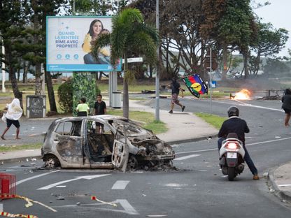 أشخاص يسيرون بجوار سيارة محترقة بينما يحمل رجل علم جبهة تحرير الكاناك الاشتراكية الوطنية في منطقة نجيا في نوميا عاصمة كاليدونيا الجديدة. 14 مايو 2024 - AFP