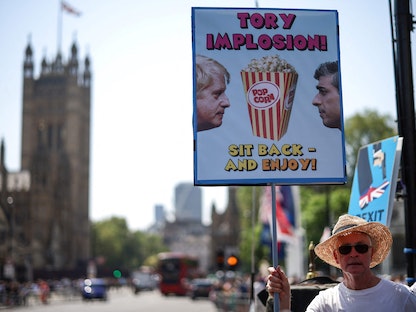 متظاهر يحمل لافتة تصور العلاقة بين رئيس الوزراء البريطاني ريشي سوناك والسابق بوريس جونسون، خلال مسيرة احتجاجية خارج مقر مجلسي البرلمان وسط لندن، 14 يونيو 2023 - AFP