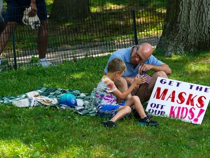 شخص يجلس مع طفل في الظل خلال مظاهرة ضد اللقاح وسط جائحة فيروس كورونا في سنترال بارك بمدينة نيويورك الأميركية، 24 يوليو 2021 - REUTERS