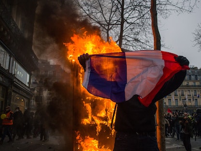 متظاهر يرفع علم فرنسا بالقرب من نيران مشتعلة في وسط العاصمة الفرنسية باريس. 23 مارس 2023 - Bloomberg