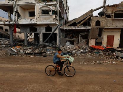 طفل فلسطيني يحمل أغطية فوق دراجة أمام منازل مُدمرة جراء غارات إسرائيلية فوق خان يونس بجنوب قطاع غزة. 27 نوفمبر 2023 - Reuters