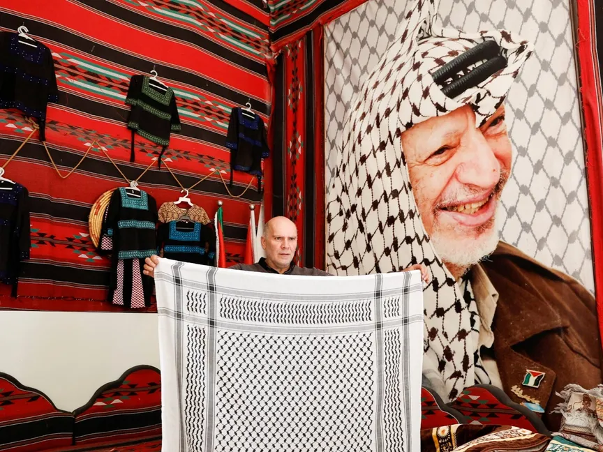 عزت حرباوي يحمل كوفية فلسطينية في مصنع نسيج أمام صورة لياسر عرفات في الخليل بالضفة الغربية. 5 ديسمبر 2023 - Reuters