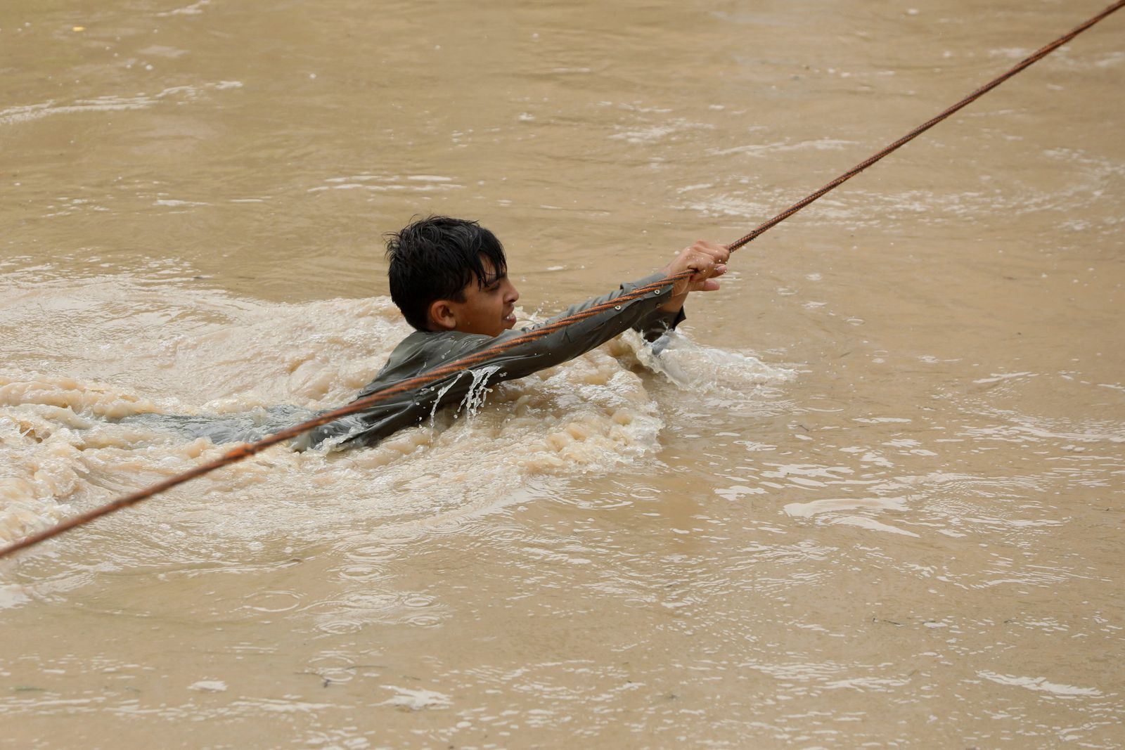 A boy crosses a flooded street, with the help of a wire fastened on both ends, following rains and floods during the monsoon season in Charsadda - REUTERS