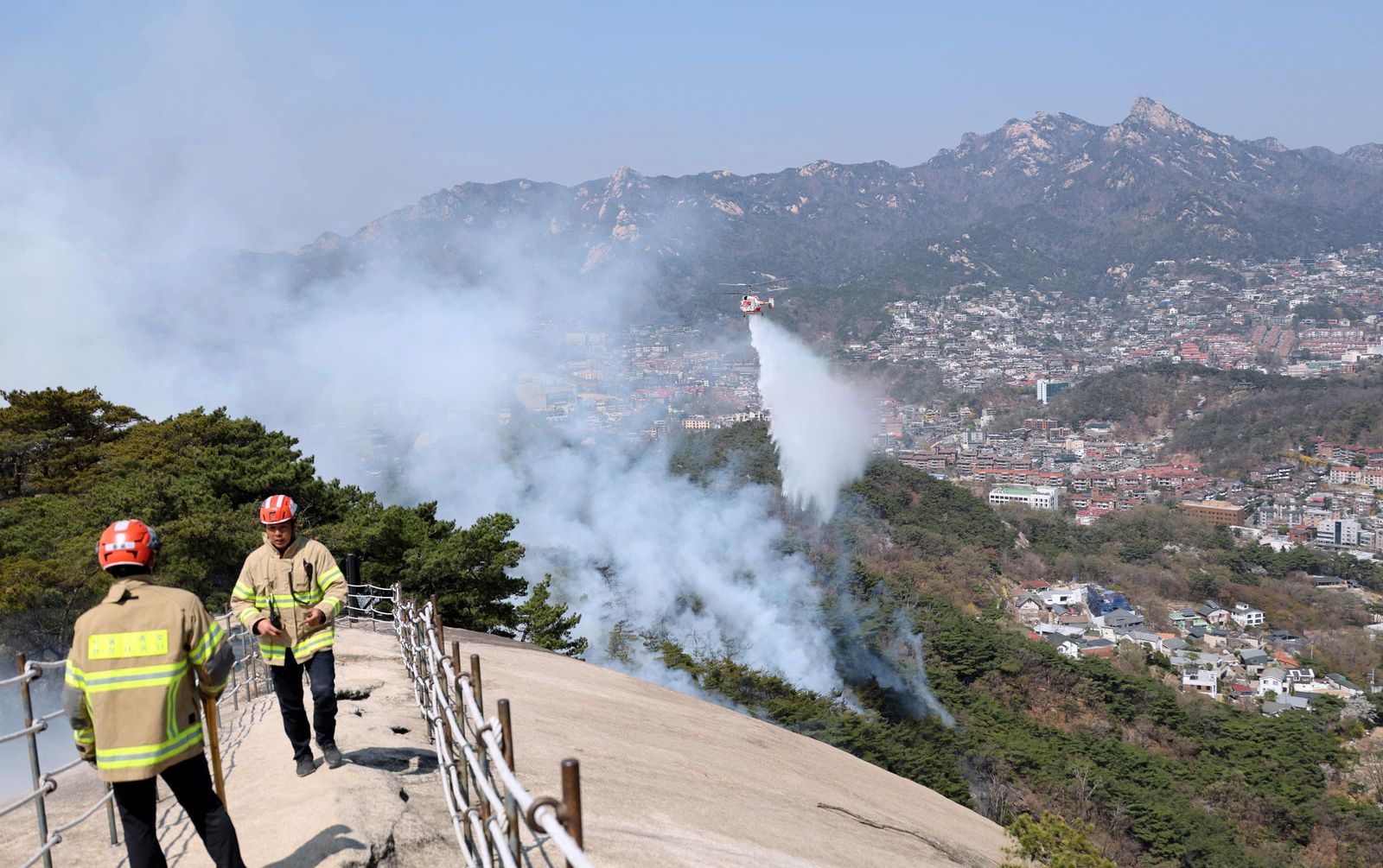 This picture taken on April 2, 2023 shows firefighters watching as a helicopter douses a wildfire on Mount Inwang in Seoul. (Photo by YONHAP / AFP) / - South Korea OUT / REPUBLIC OF KOREA OUT  NO ARCHIVES  RESTRICTED TO SUBSCRIPTION USE - AFP