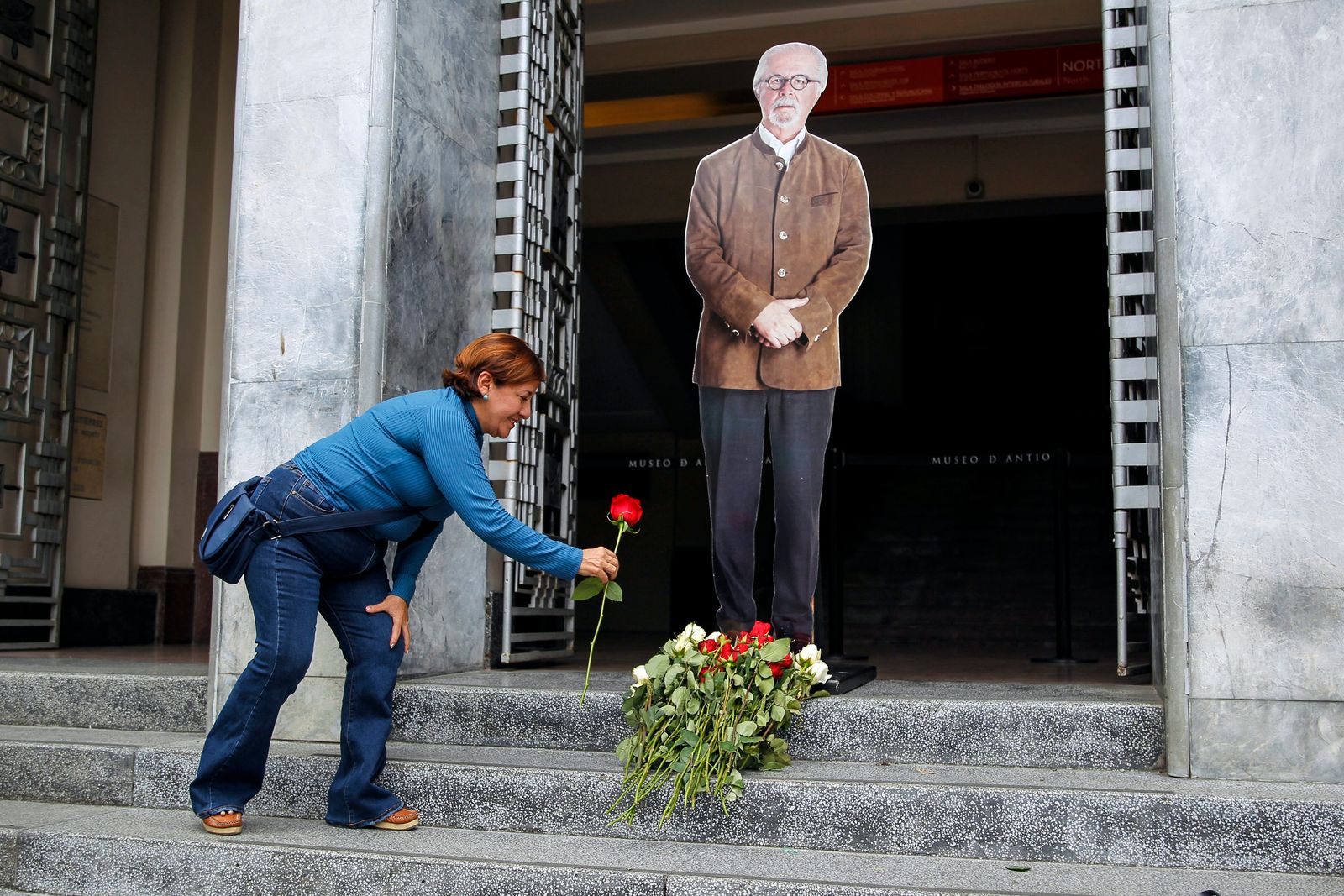 A woman lays a red rose at the feet of an image of Colombian artist Fernando Botero, at the entrance of the Antioquia Museum in Medellin, Colombia, on September 15, 2023, on the day of his passing. - Colombia's most famous artist, Fernando Botero, who was known for his voluptuous depictions of people and animals, has died aged 91, President Gustavo Petro announced on September 15, 2023. Botero's works of plump and slightly surreal forms -- often with a touch of satire or political commentary -- are enormously popular and have been displayed in cities around the world, including Bogota, Madrid, Paris, Singapore and Venice. (Photo by Fredy BUILES / AFP) - AFP