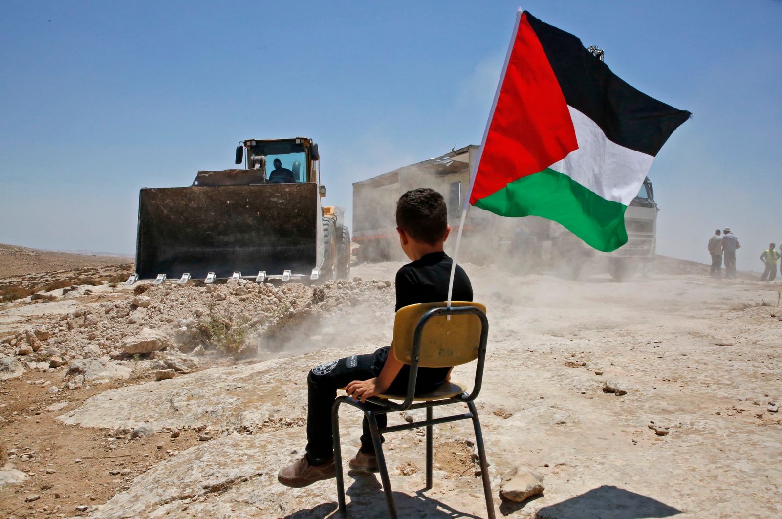 صبي فلسطيني يجلس على كرسي ويحمل علم فلسطين بينما تهدم السلطات الإسرائيلية مدرسة في قرية يطا ، جنوب مدينة الخليل بالضفة الغربية المحتلة - AFP