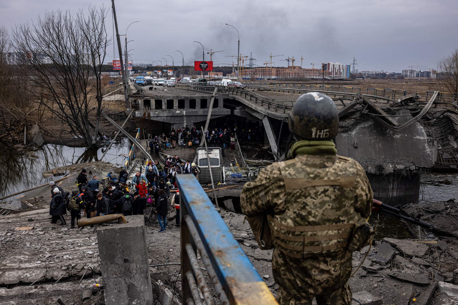 A Ukrainian serviceman looks on as evacuees cross a destroyed bridge as they flee the city of Irpin, northwest of Kyiv, on March 7, 2022. - Ukraine dismissed Moscow's offer to set up humanitarian corridors from several bombarded cities on Monday after it emerged some routes would lead refugees into Russia or Belarus. The Russian proposal of safe passage from Kharkiv, Kyiv, Mariupol and Sumy had come after terrified Ukrainian civilians came under fire in previous ceasefire attempts. (Photo by Dimitar DILKOFF / AFP) - AFP