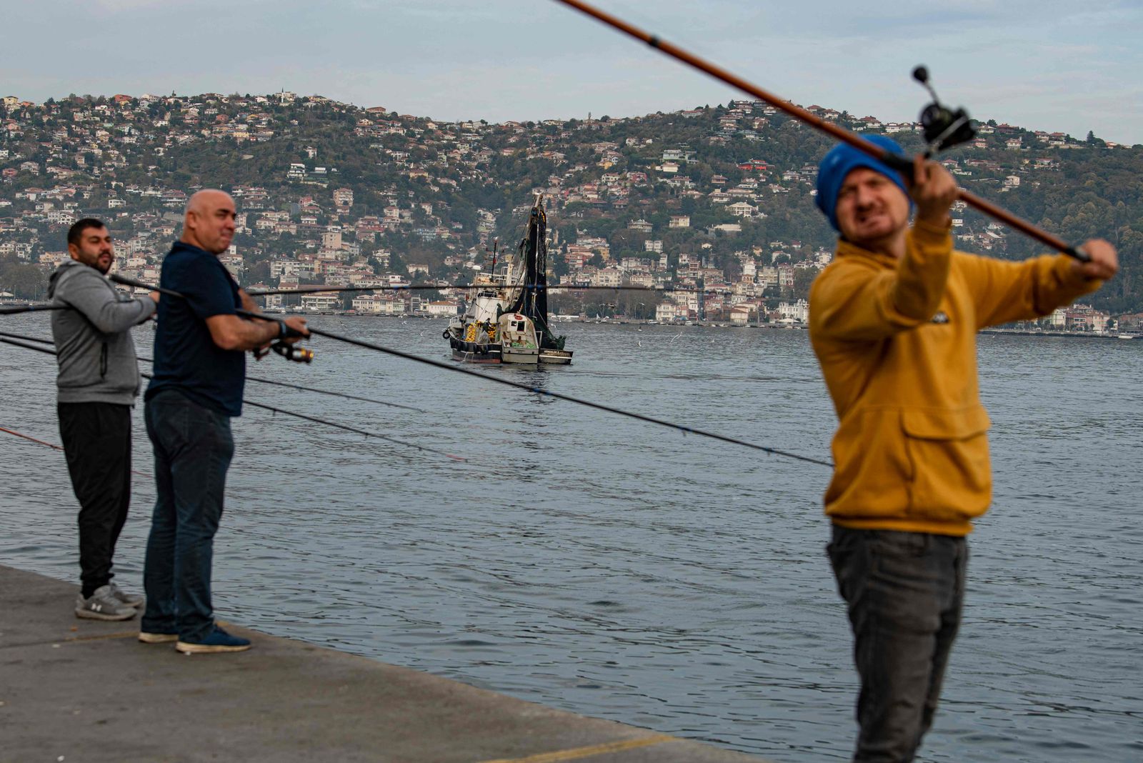 Anglers fish on the bank of the Bosphorus as a fishing boat lays its nets in the Marmara sea off the cost of Istanbul, on November 6, 2022. - It is high season for the popular variety of tuna, with shoals teeming through the Bosphorus on their way from the Black Sea to the Mediterranean. But pulled taut across the strait are fishing nets more than a kilometre (3,280 feet) long. Anglers who cram shoulder to shoulder along the banks say the nets leave them with little chance, and the fish with even less. (Photo by Yasin AKGUL / AFP) - AFP