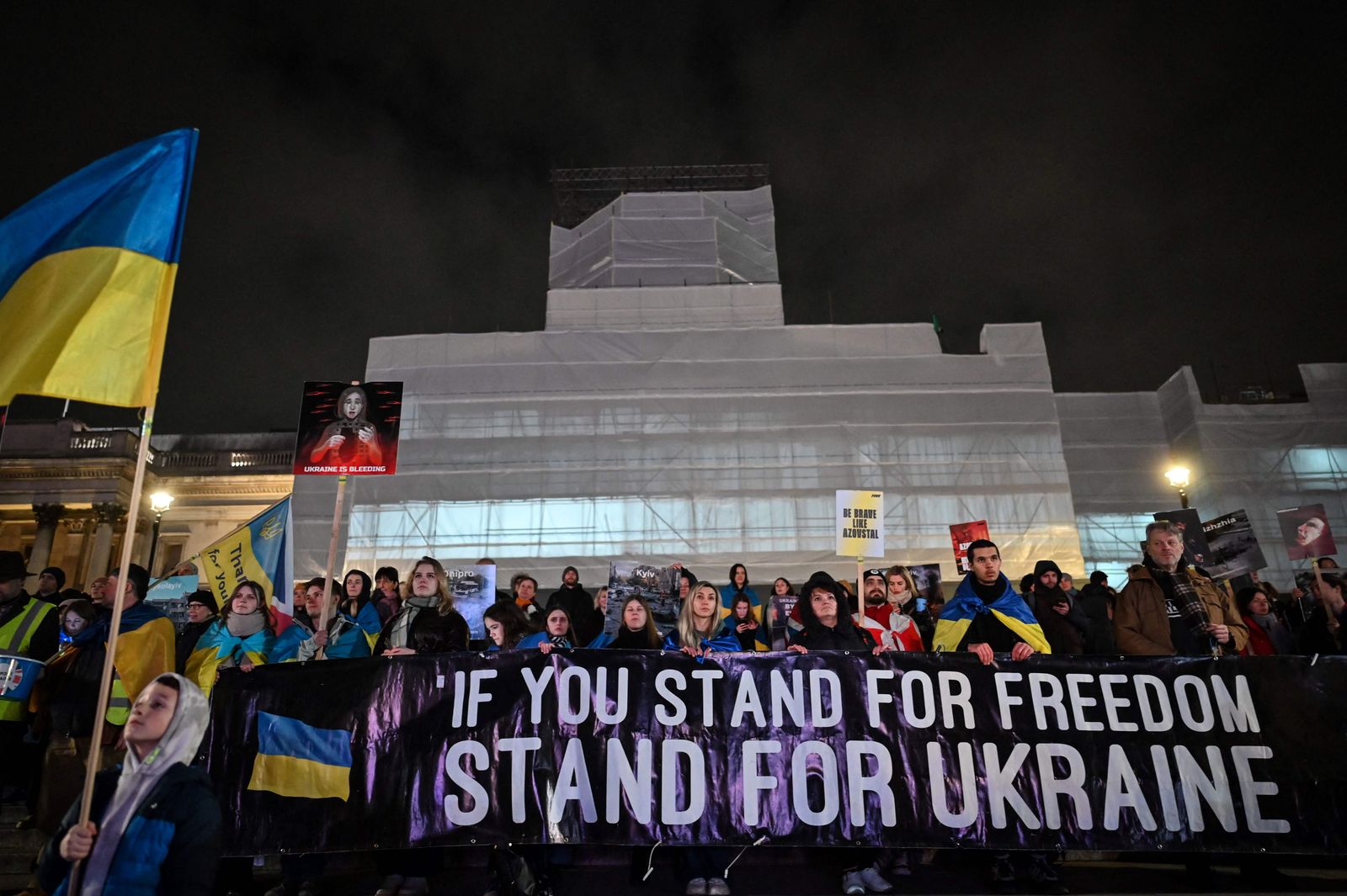 أشخاص يلتفون بالأعلام الأوكرانية ويحملون لافتات خلال وقفة احتجاجية بميدان ترافالجار وسط لندن. 23 فبراير 2023 - AFP
