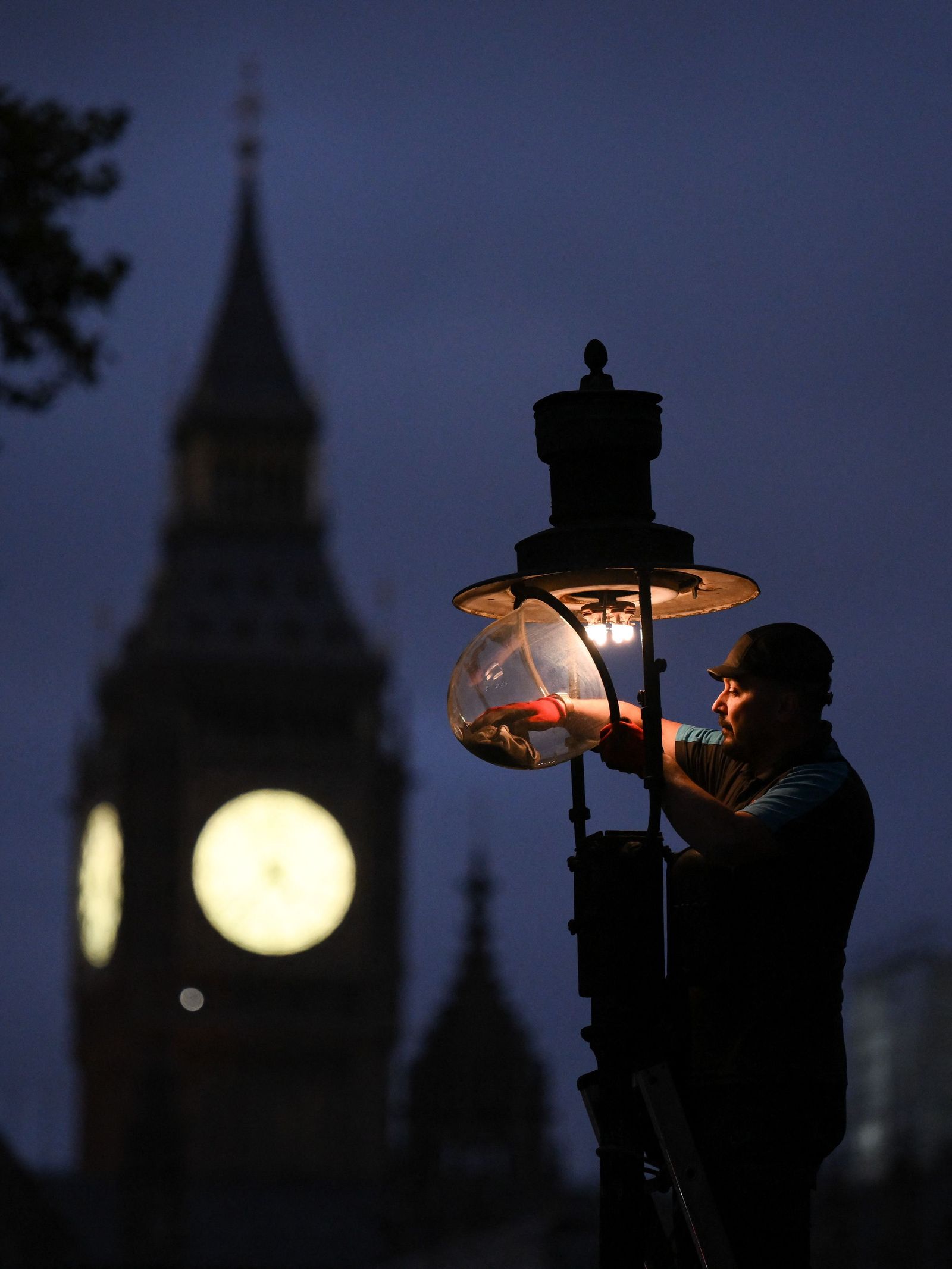 A British Gas engineer services a gas-powered lamp near the Houses of Parliament in central london on November 11, 2022. - Intrigued tourists watch as Paul Doy climbs a ladder outside London's Westminster Abbey and lifts the globe of a gas street lamp. Winding its timer, he then ignites a small cloth mesh, creating a distinctive soft warm light that illuminates the darkness. 