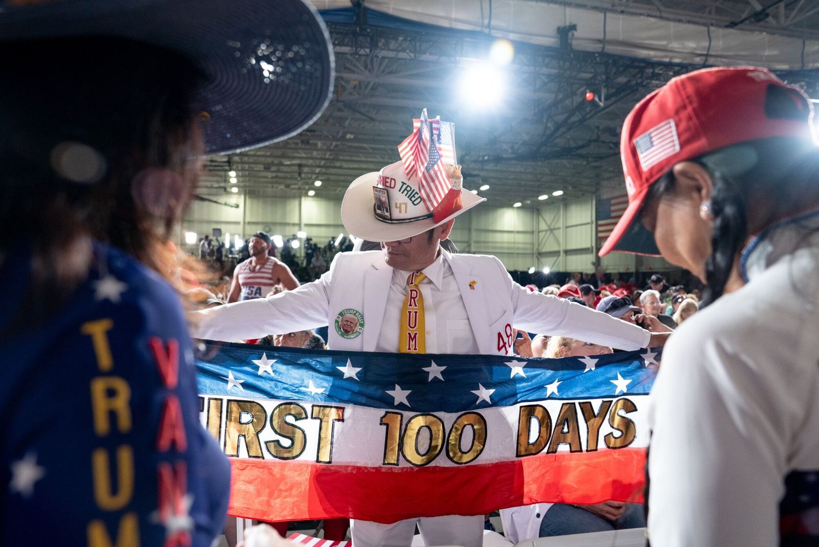 An attendee holds a 'First 100 Days' banner prior to a 100th day rally with US President Donald Trump, not pictured, in Warren, Michigan, US, on Tuesday, April 29, 2025. Trump's aggressive pace in the first 100 days of his second term is accompanied by greater confidence in his political instincts and an inner circle even more stocked than last time with loyalists. Photographer: Sarah Rice/Bloomberg