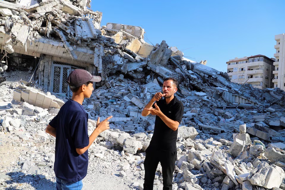 المؤثر الفلسطيني الأصم باسم الحبل يقف مع شخص آخر يترجم لغة إشارته، وخلفهما منازل مدمرة، مدينة غزة، 5 أغسطس 2025