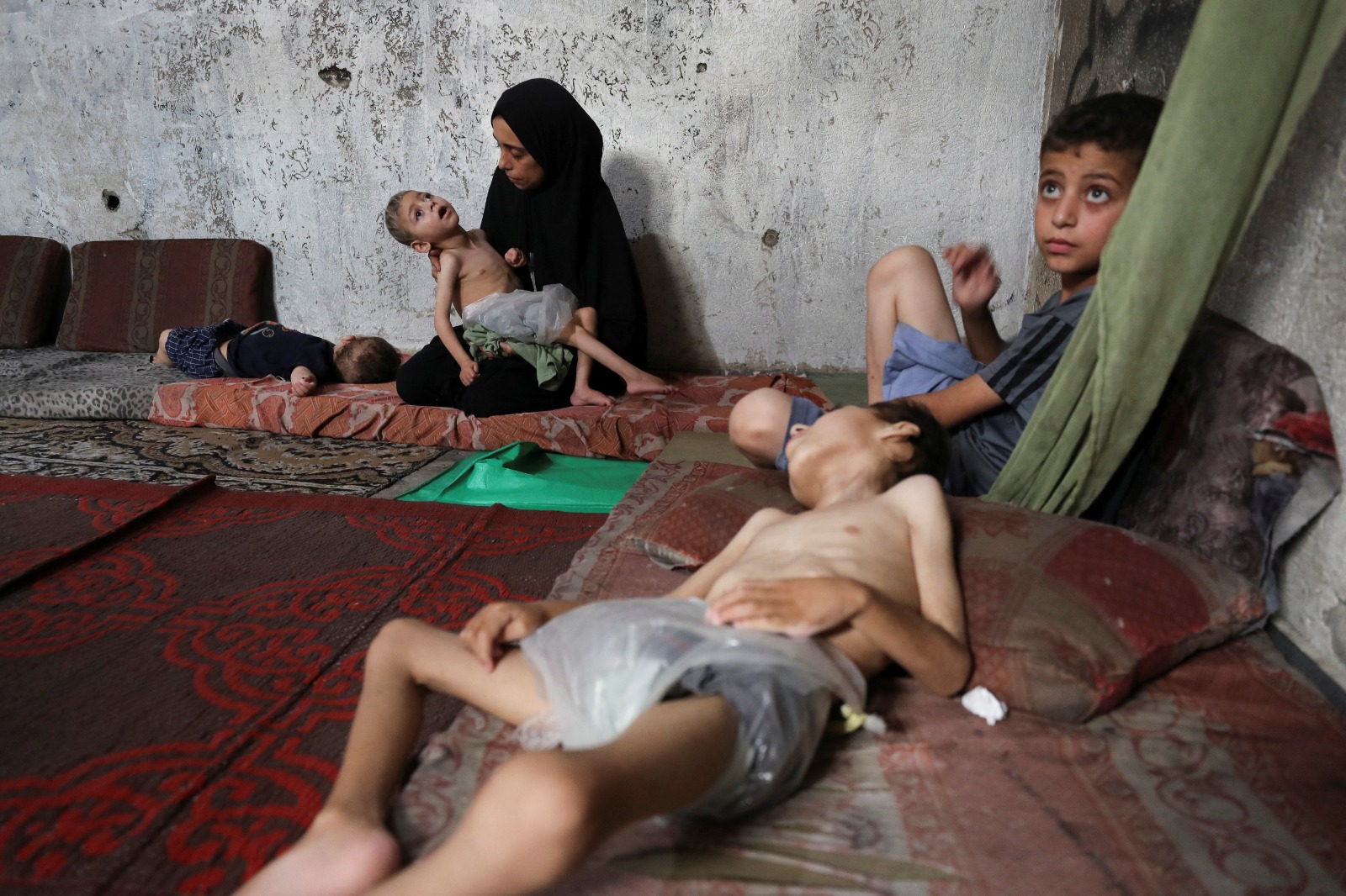 أم فلسطينية تحمل ابنها الذي يعاني من سوء التغذية ويعاني من شلل دماغي على فراش في مدرسة وسط أزمة الجوع في قطاع غزة. 24 يوليو 2025