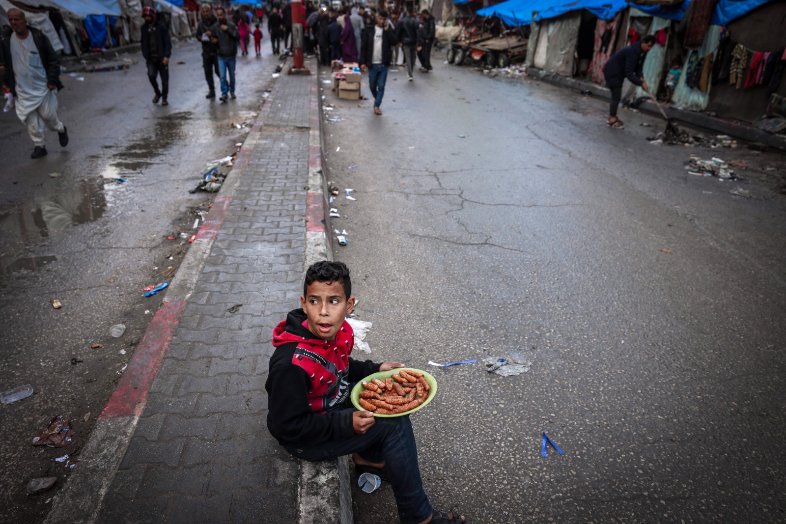 طفل نازح فلسطينيي يبيع الحلوى ينتظر الزبائن على رصيف شارع بالقرب من مخيمات للنازحين في رفح بأول أيام عيد الفطر. قطاع غزة، فلسطين. 10 أبريل 2024